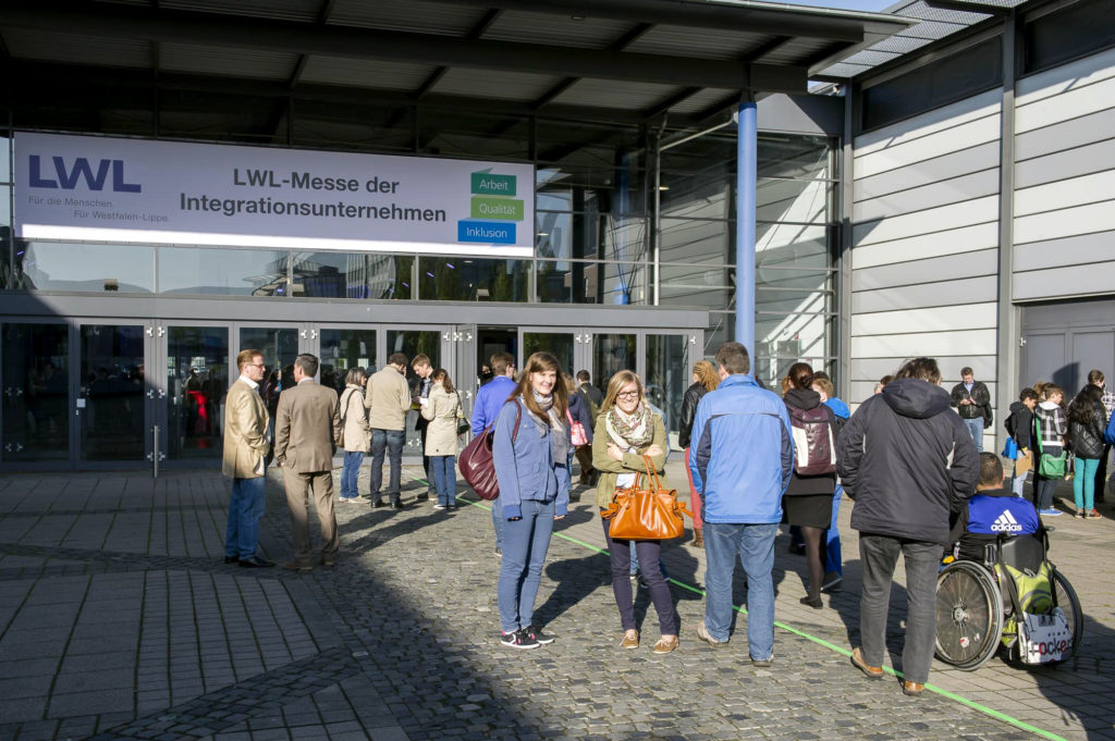 Eine Gruppe Menschen, einer davon im Rollstuhl, in der Sonne vor dem Eingang zur LWL-Messe 2014.