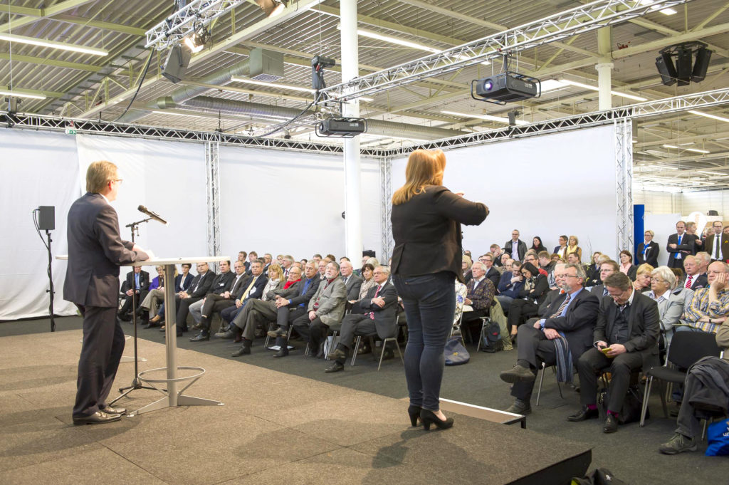 Auf der Hauptbühne spricht ein Mann vor sitzendem Publikum. Eine Gebärdendolmetscherin übersetzt simultan.