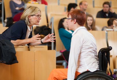 Eine Professorin tauscht sich am Podium in einem vollen Vorlesungssaal mit einer Bildungsfachkraft im Rollstuhl aus.
