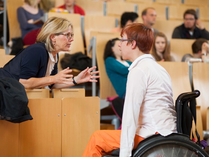 Eine Professorin tauscht sich am Podium in einem vollen Vorlesungssaal mit einer Bildungsfachkraft im Rollstuhl aus.