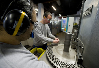 Zwei Mitarbeiter, die einen Ohrschutz tragen, sortieren Metallfedern in eine Schleifmaschine.