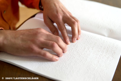 Auf dem Foto sind zwei Hände zu sehen, die ein Heft mit Brailleschrift abtasten.