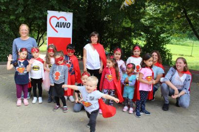 Kinder mit Superhelden-Kostümen haben sich draußen zum Gruppenfoto aufgestellt, vorne tanzt ein Kind mit rotem Umhang aus der Reihe