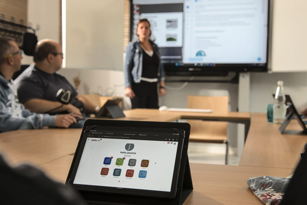Ein Tablet mit geöffneter Lwerk-App steht auf einem Tisch in einem Besprechungsraum, im Hintergrund eine stehende Person vor einem Bildschirm und zwei sitzende Personen daneben.