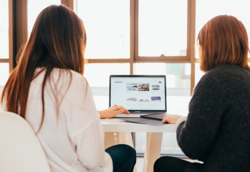 Zwei Frauen sitzen vor einem Laptop, die Frauen sind von hinten zu sehen
