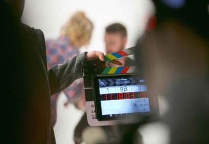 Blick auf eine Filmklappe, dahinter eine Filmszene mit zwei Menschen vor einem hellen Hintergrund.