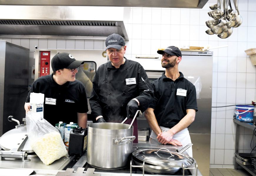 enedikt Minnig, Frank Kölling und Aqid Ibrahim (von links) stehen in Arbeitskleidung in der Großküche des Unternehmens hinter einem großen Kochtopf, Frank Kölling rührt, die drei sprechen lachend miteinander.