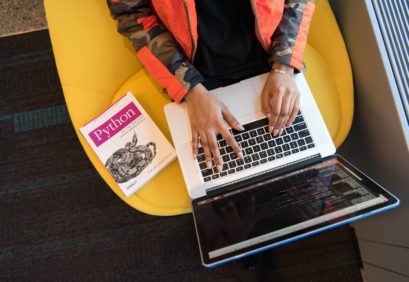 Die Hände einer Frau auf der Tastatur eines Laptops von oben, neben ihr liegt ein Buch zu einer Programmiersprache.