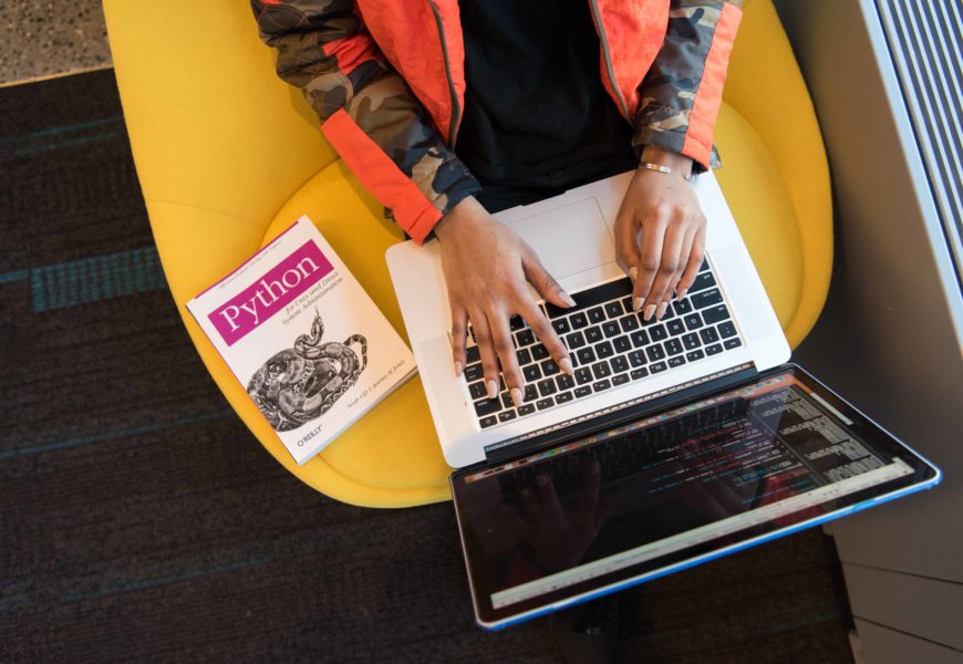 Die Hände einer Frau auf der Tastatur eines Laptops von oben, neben ihr liegt ein Buch zu einer Programmiersprache.