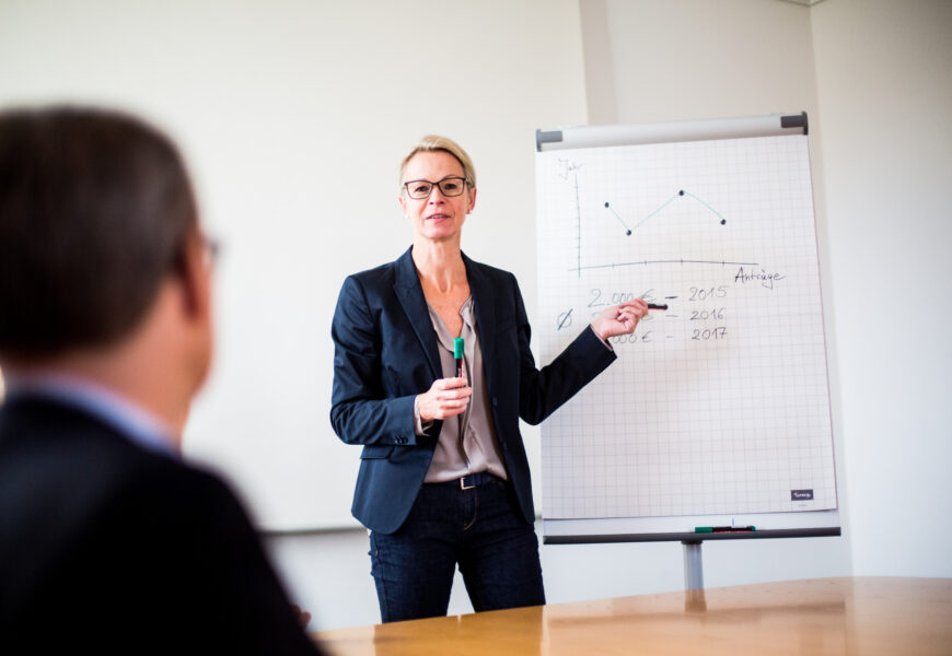 An einem Flipchart stehende Seminarleiterin erklärt den Anwesenden etwas