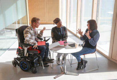 Drei Personen sitzen an einem runden Tisch in einem hellen Raum, eine davon im Elektrorollstuhl; eine Person gebärdet, die anderen hören zu.