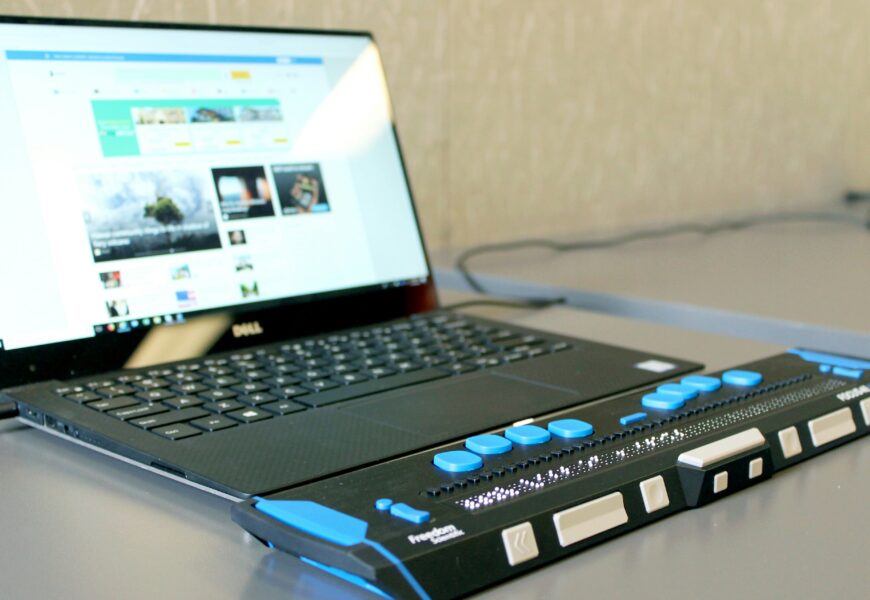 Ein aufgeklappter Laptop steht auf einem Tisch. Davor liegt ein elektronisches Braillezeilen-Gerät mit blauen und weißen Tasten sowie einer länglichen Braille-Anzeige.
