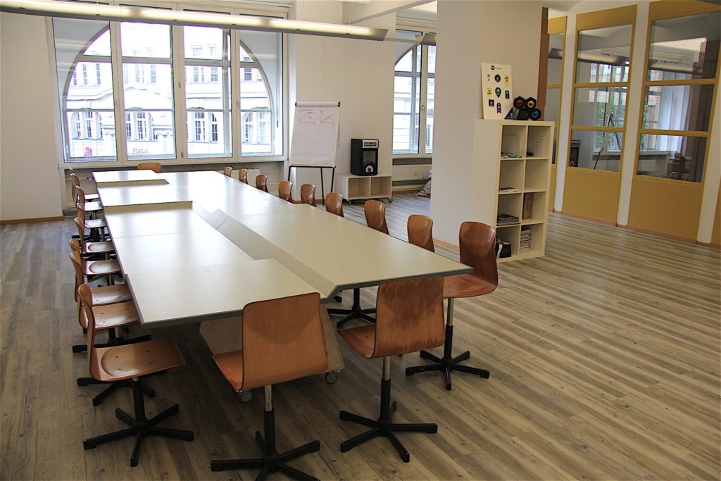 Ein großer, leerer Tisch, dessen Tischplatte verschiedene Höhen hat. Er steht in den Räumen des Coworking-Spaces in Berlin. Drumherum stehen viele Stühle.