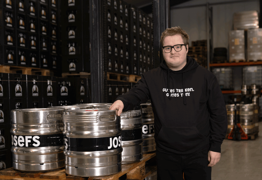 Nico Wichmann in schwarzem Pulli lehnt im Lager der Josefsbrauerei an silbernen Bier-Fässern.