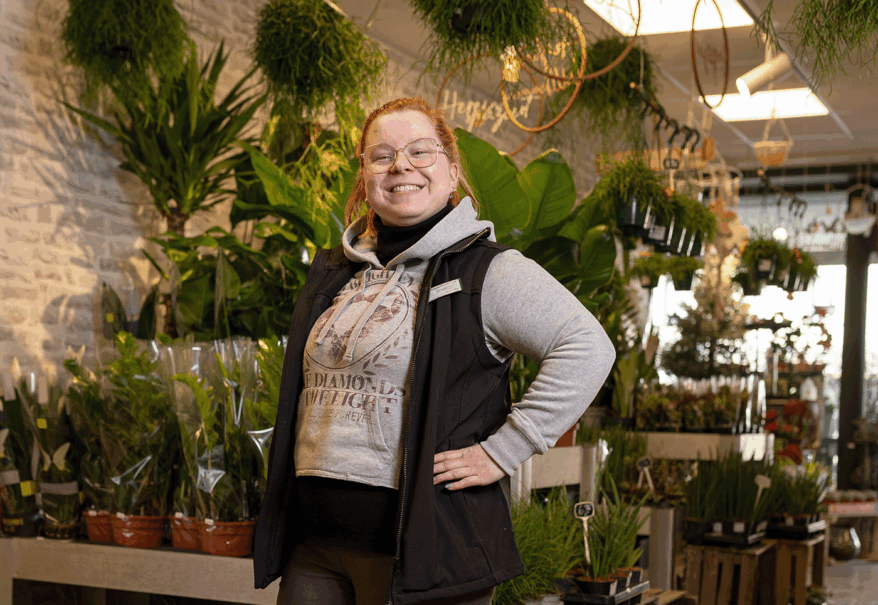 Denise Urban in grauem Pulli und Weste steht mitten in einem Blumenladen und stützt die Hände in die Hüften.