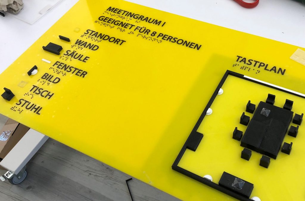 Auf einer rechteckigen, grellgelben Platte ist rechts ein Plan des Coworking-Spaces mit Miniaturmöbeln nachgebildet. Links sind die einzelnen Möbel noch einmal einzeln aufgelistet und werden mit Laut- und Blindenschrift erklärt.