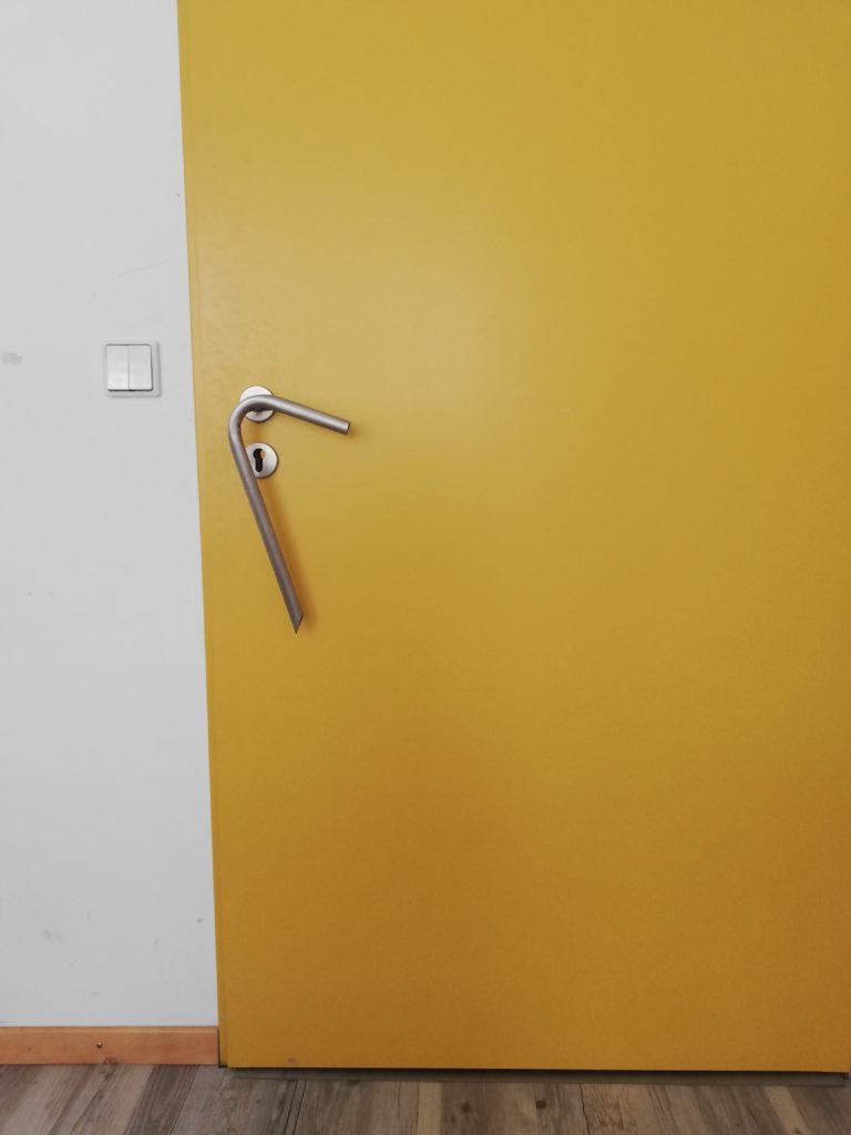Eine gelbe Tür mit einer nach unten langgezogenen Klinke, die auch kleine Menschen gut erreichen können.
