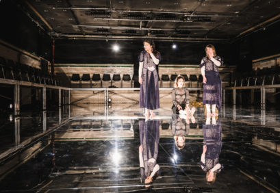 Drei Schauspielerinnen auf einer spiegelnden Bühne (zwei stehen außen, eine sitzt mit ausgestreckten Beinen in der Mitte der beiden anderen), die mit Körpersprache und Mimik etwas ausdrücken.